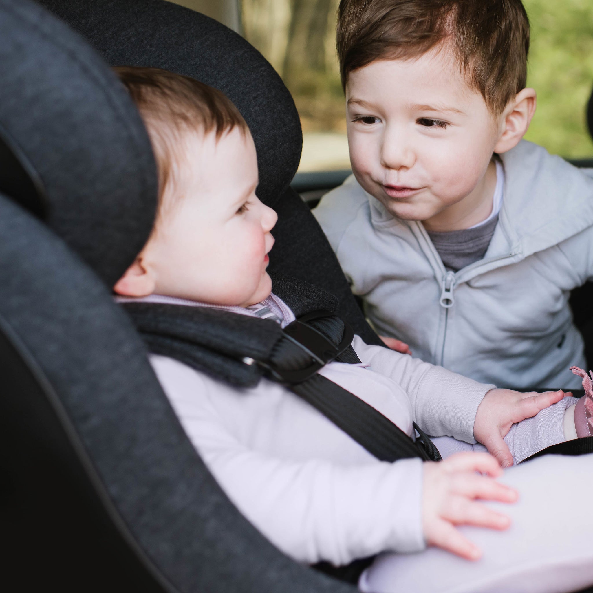 Two children sitting close together, one in a car seat all-groups