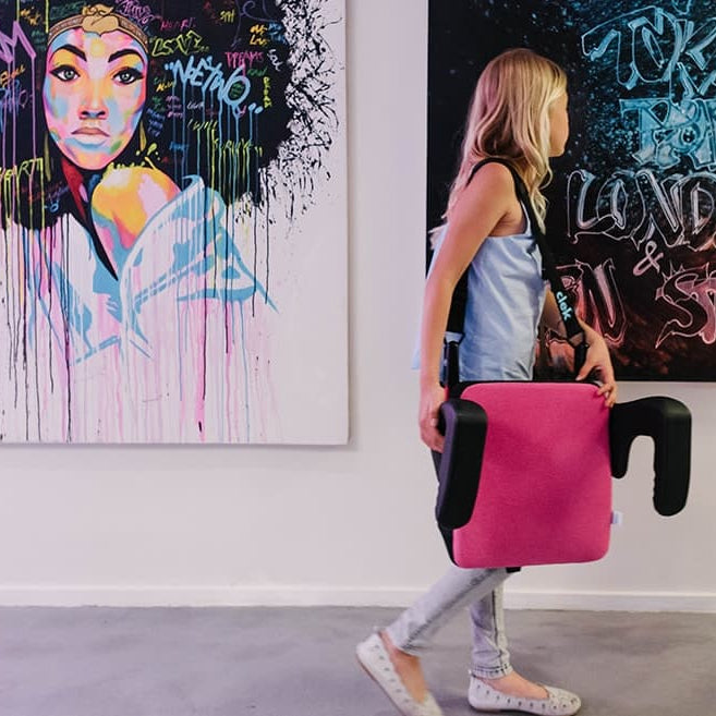 Young girl carrying her Clek olli booster seat for kids in flamingo fabric using the strap-thingy accessory