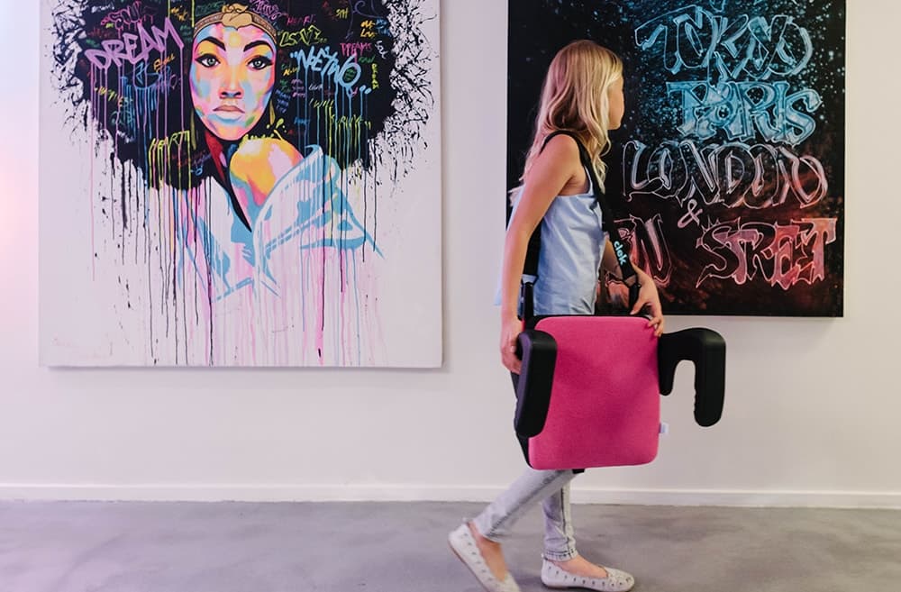 Young girl carrying her Clek olli booster seat for kids in flamingo fabric using the strap-thingy accessory